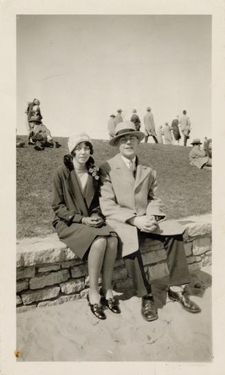 A photograph of two people sitting on a stone ledge in front of a small grass hill. There are o ...