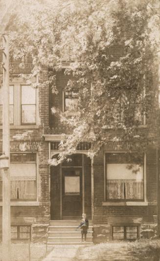 Black and white photograph of a boy standing on the steps of a three story, brick house.