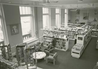 Picture of interior view of library branch showing shelves and chairs and people standing and s ...