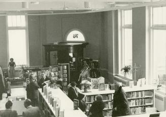 Picture of interior view of library branch showing crowd of people standing amongst shelves. 
