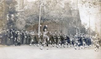 A photograph of a military parade, with dozens of men in kilts and hats carrying and playing ba ...