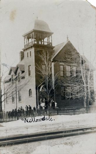 Black and white photograph of a two story school building with a turret. Children are standing  ...