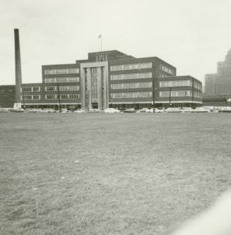 A photograph of a six story office building, with a grass lawn and parking lot in front of it.  ...