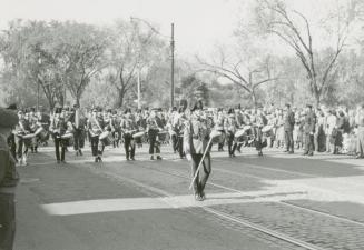 A photograph of a band marching down a paved city stret, with spectators and police officers wa…