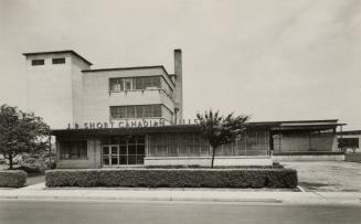 Black and white photo postcard depicting the exterior front of a building, with a sign on top o ...