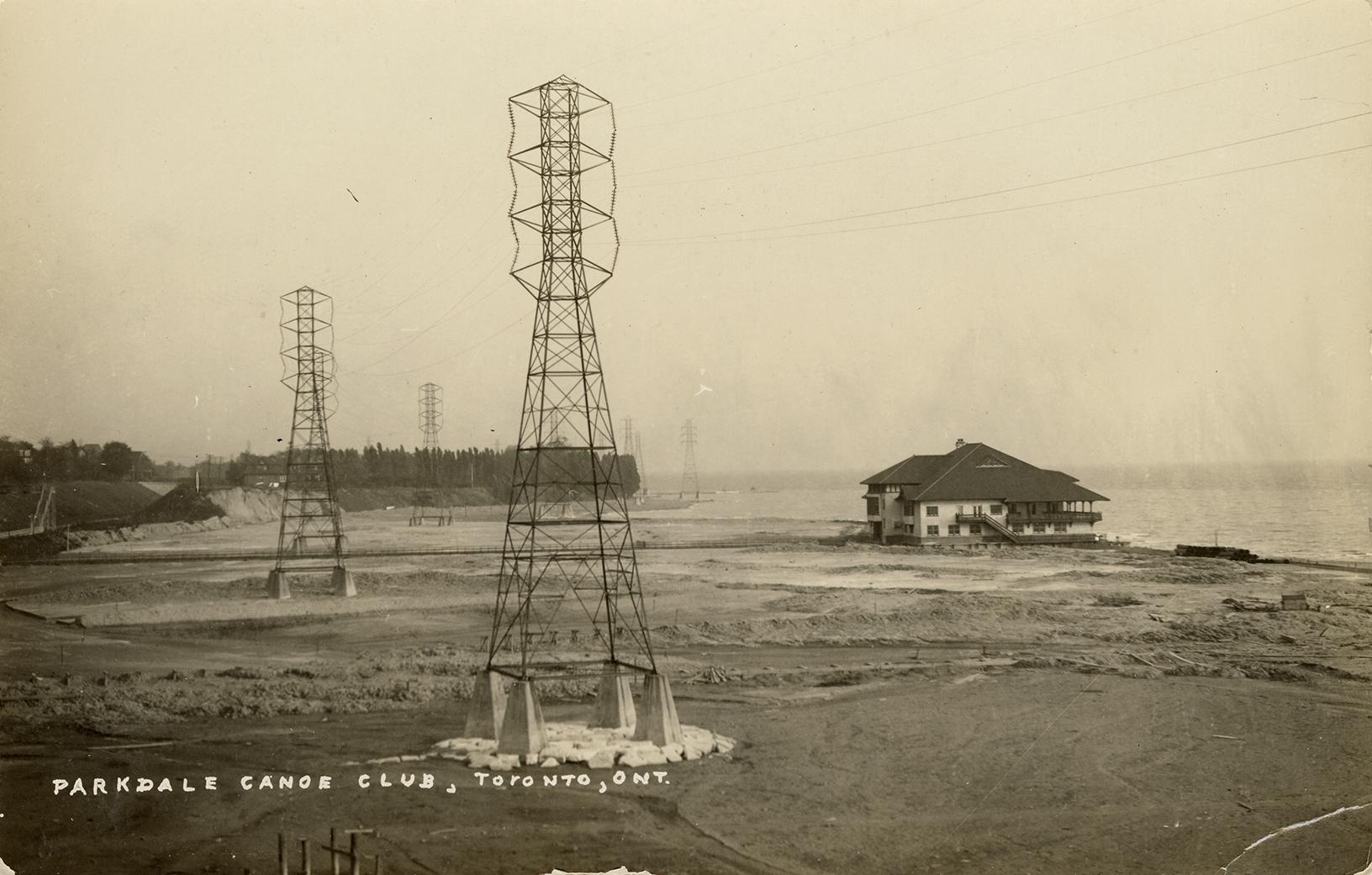 Black and white photograph of a large building built on a shoreline. Electrical transmission to ...