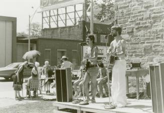 Picture of two men playing guitars on a stage with a few people at the side looking at things o ...