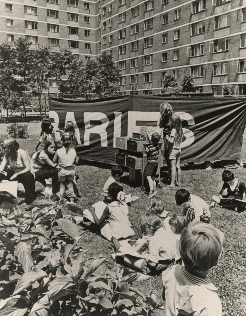 Picture of a group of children on a lawn looking at books with apartment building in background ...