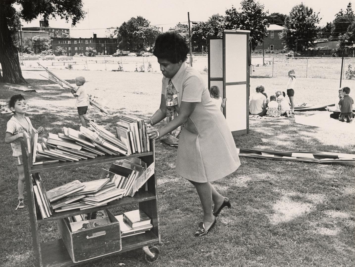 Picture of a group of children on a lawn looking at books with apartment building in background ...