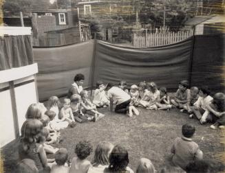 Picture of a group of children in a park listening to a story from a librarian. 