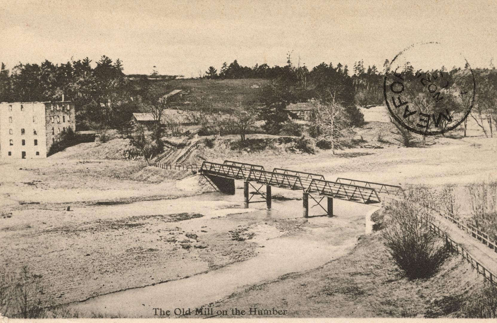 Black and white photograph of a river running under a bridge beside a square, stone building.