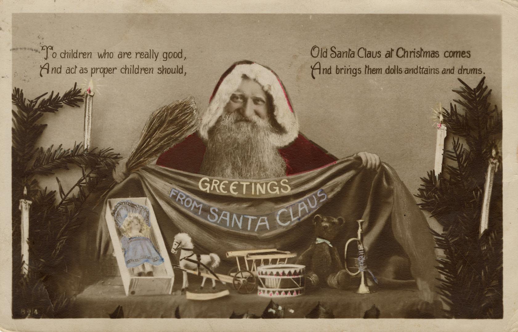 Sepia toned photograph of Santa standing behind a table with toys on it. Greenery with candles  ...