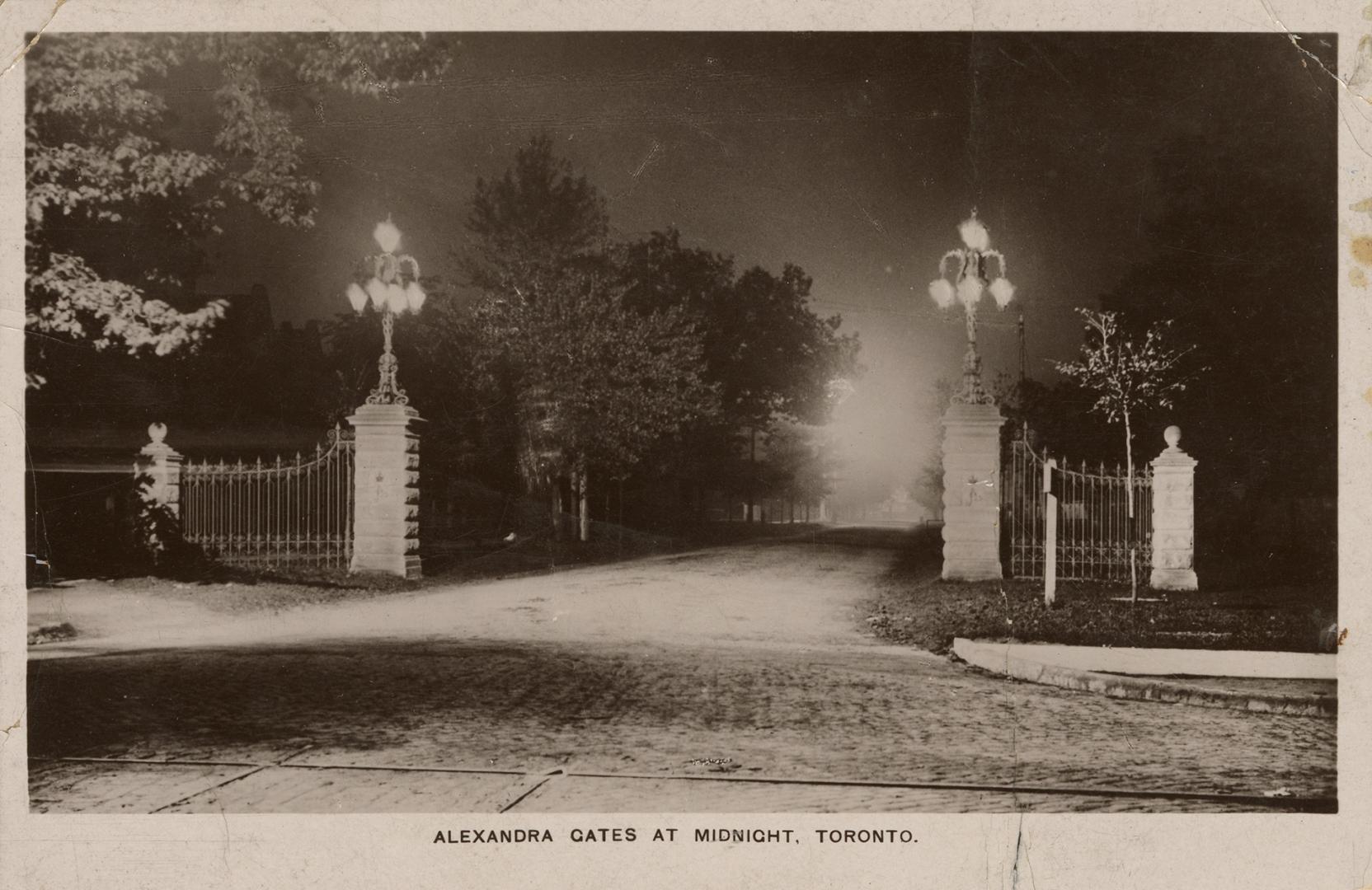 Black and white photograph of an open gate with a wide road passing through, taken at night.