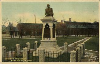 Colour photo postcard depicting a monument with a bust at the top, surrounded by a fenced-off a ...