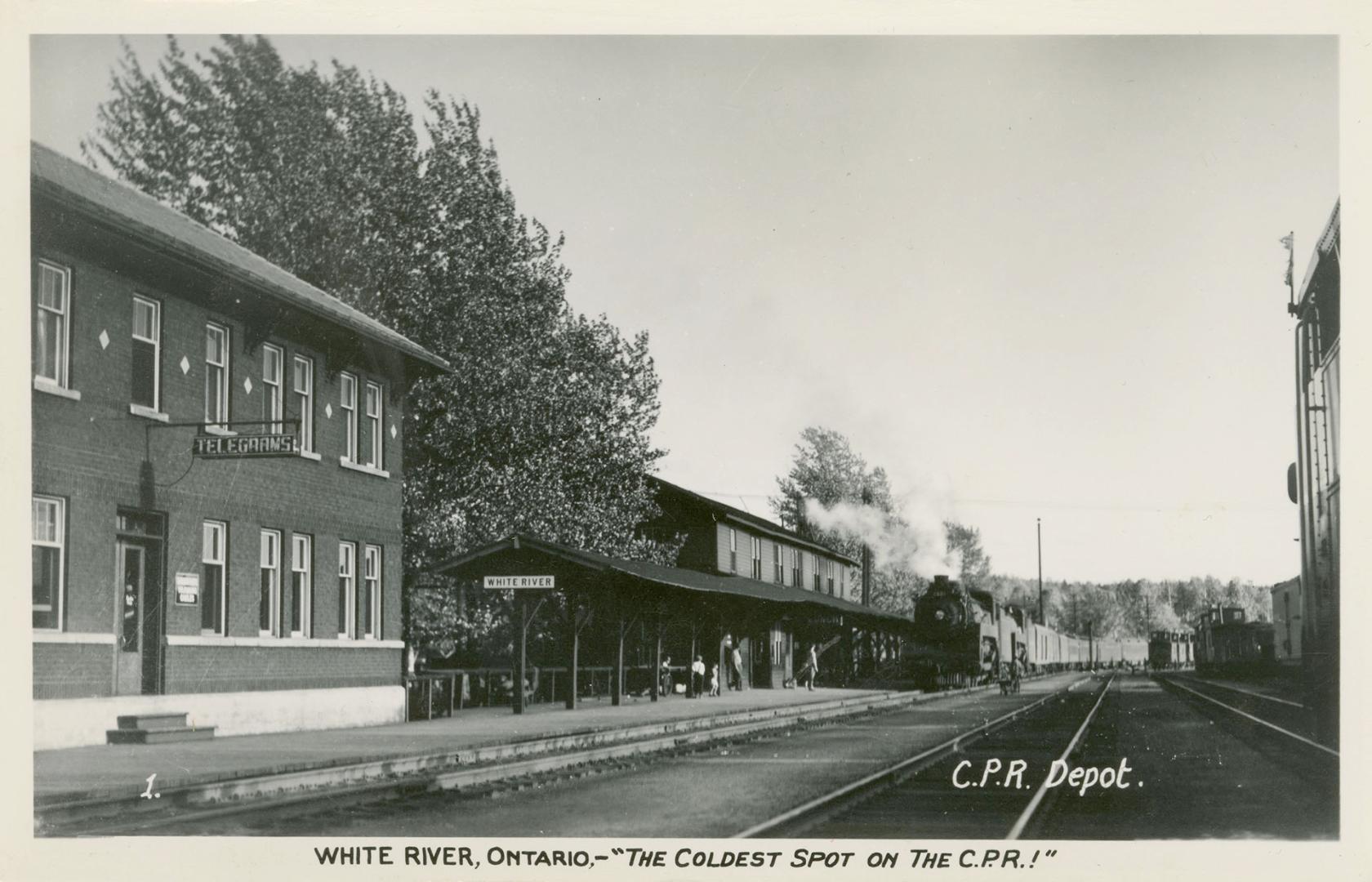 Black and white photograph of rail road tracks running past a railway station.