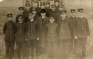 Black and white photo postcard depicting a group of 16 men in uniform with caps. A man in the c ...