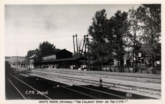 Black and white photograph of rail road tracks running past a railway station.