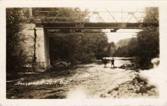 Photograph of two people standing on the bank of a river underneath a bridge