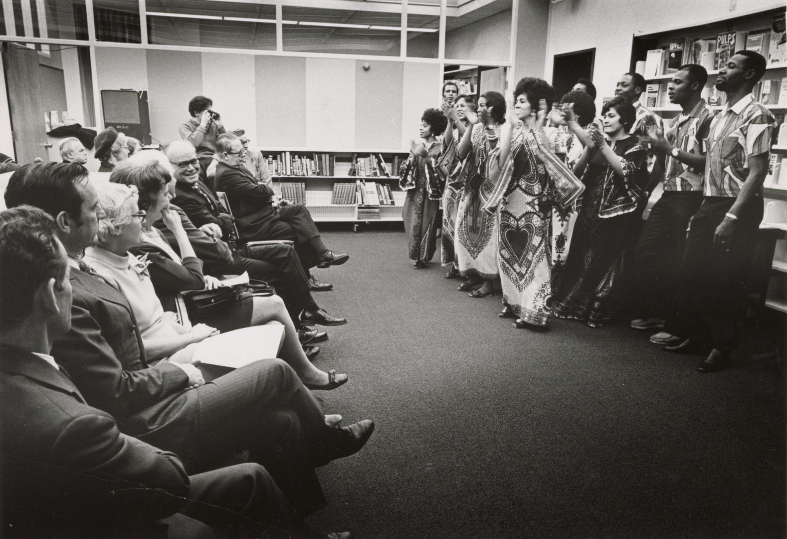 Group of singers perform in front of seated crowd in a library. 