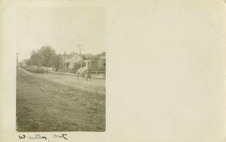 Black and white postcard depicting an inset photo of a dirt road with many men trailing behind  ...