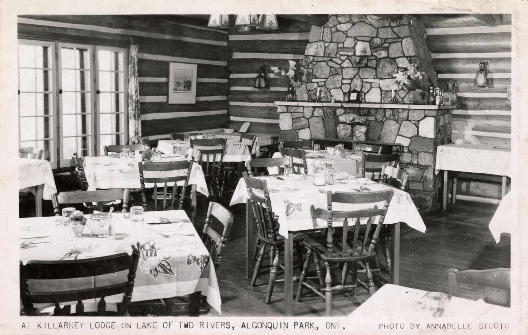 Black and white photograph of tables set for four people in a room in log cabin with fieldstone ...