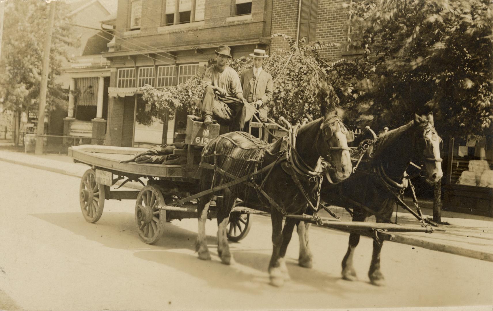 Sepia-toned photo postcard depicting two-men aboard a two-horse-drawn wagon. The back of the ca ...