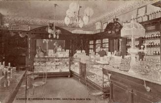 Brown-toned photo postcard depicting the interior of a shop with a man standing in front of a c ...