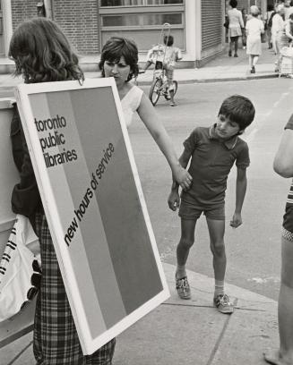 Person wearing a sandwich board advertising library hours stands on the street with boy looking ...