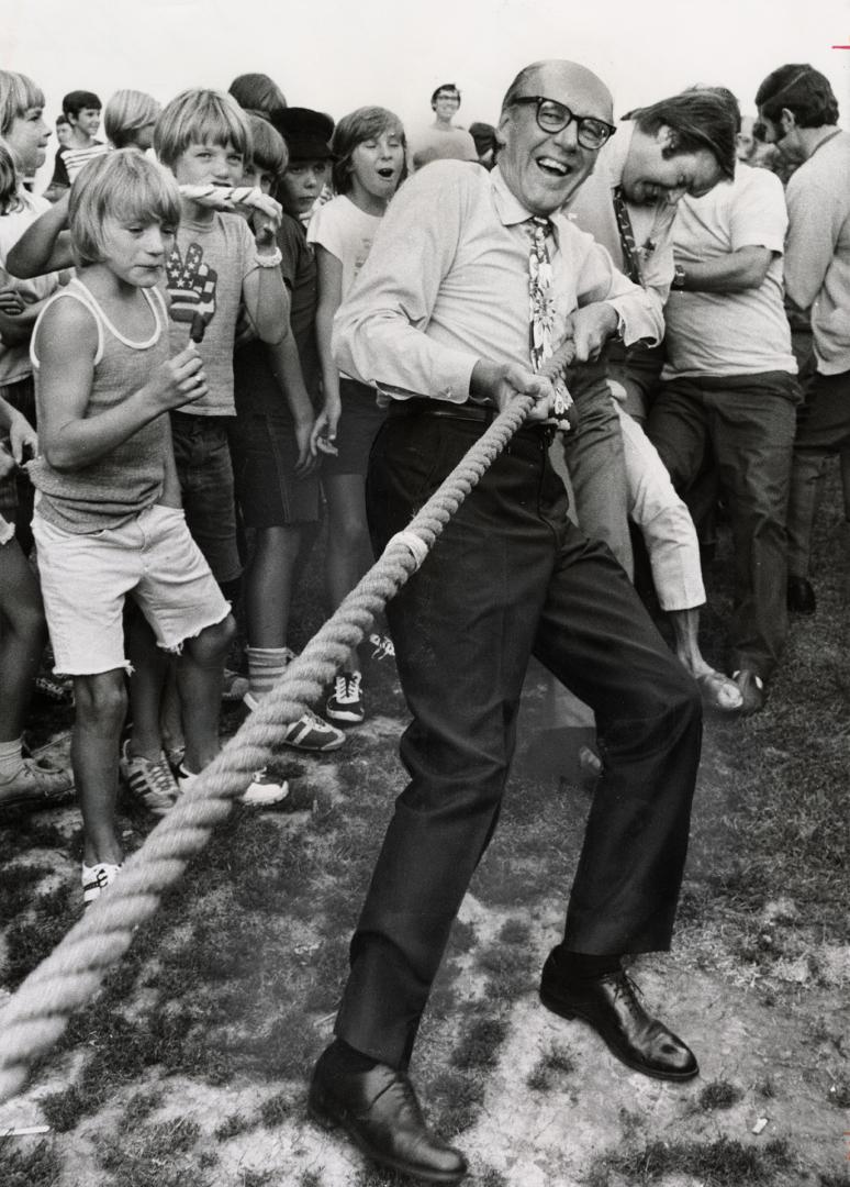 Older white man leaning back tugs on a thick rope surrounded by a large group of boys