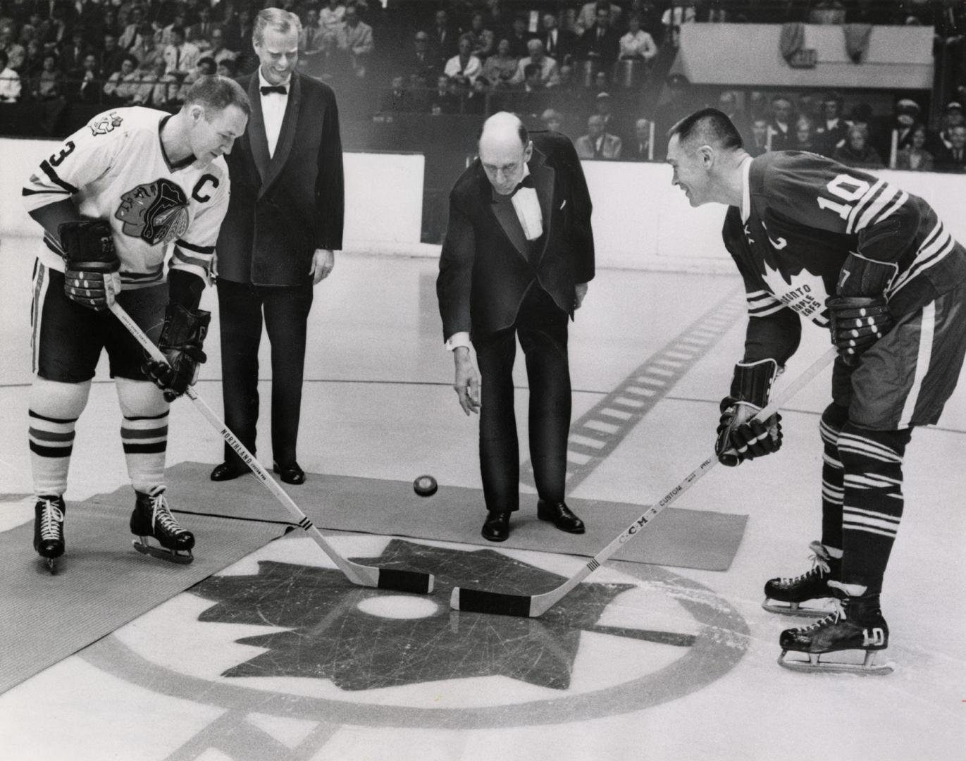 Bald white man in tuxedo drops puck between two hockey players at centre ice. A blond man in a  ...