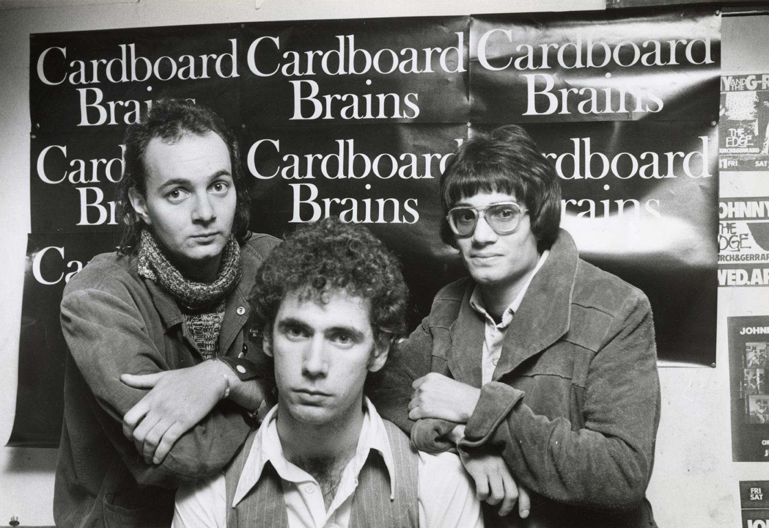 Three young stand before a poster with the text, " Cardboard Brains" repeated on it. 