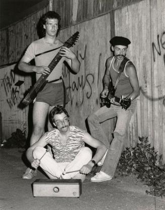 Three young men dressed casually in front of tall wooden fence with graffiti. One sits on the g…