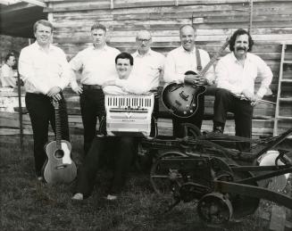 6 men in white shirts and dark pants stand in front of a weather beaten wooden wall with an old…