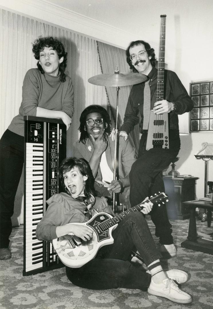 Two young women and two young men in a cluster in a living room setting with a bass guitar, gui…