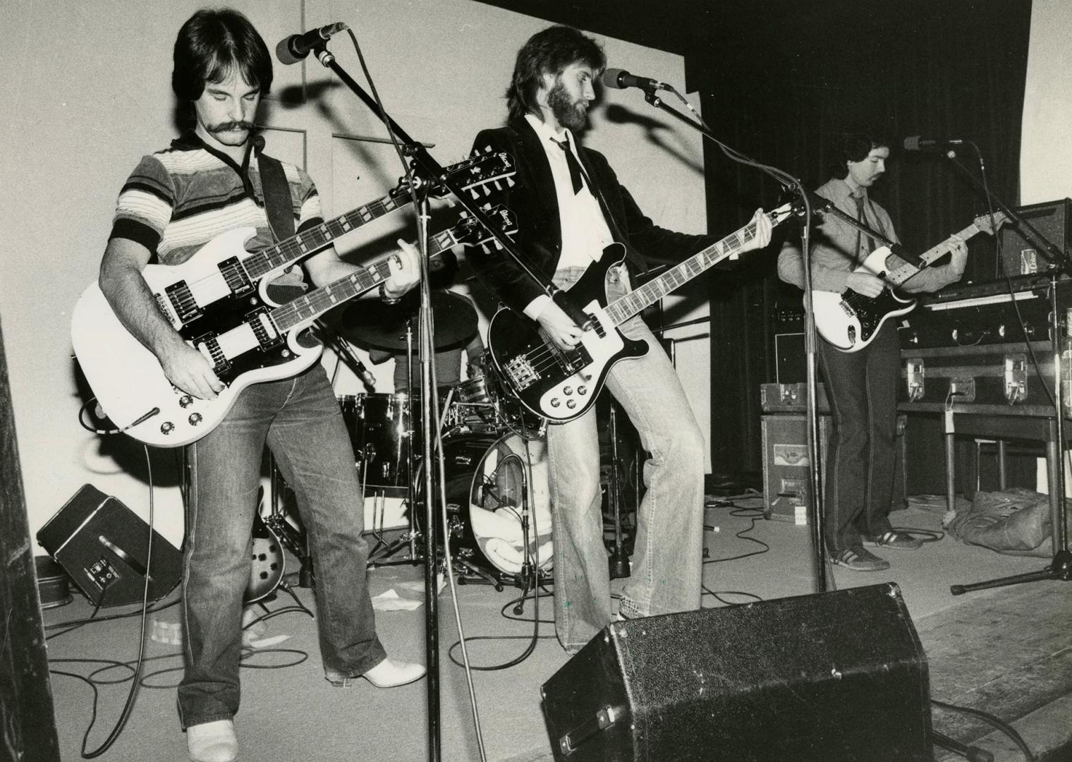 3 young men on a bare stage in front of drum kit, each has a guitar and is standing behind micr…