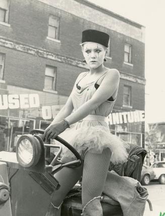 Young woman in mime costume and makeup standing up in a jeep in front of old 3 story storefront…