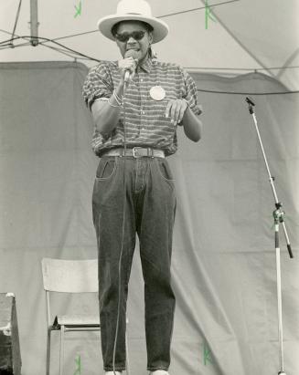 Full body shot. Man in sun glasses and sun hat sings into hand held microphone. 