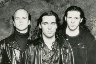 Head and shoulders, three young men with long hair in black jackets. Mottled wallpaper backgrou…