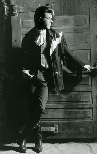 Full body shot of young man looking right wearing jeans, cowboy boots and a country-western bla…