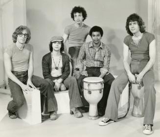 five young men sitting in row on boxes and tabla drums on a light solid colour background. 