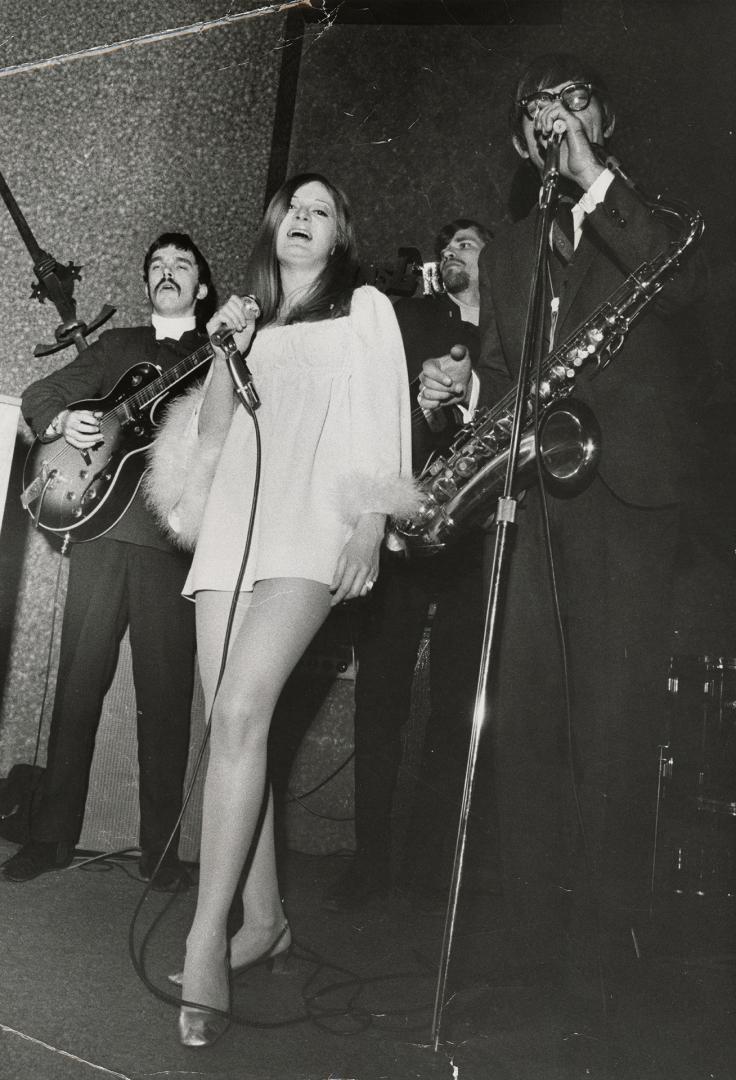 On darkened stage, young woman in white mini dress singing with microphone. Beside her, a young…