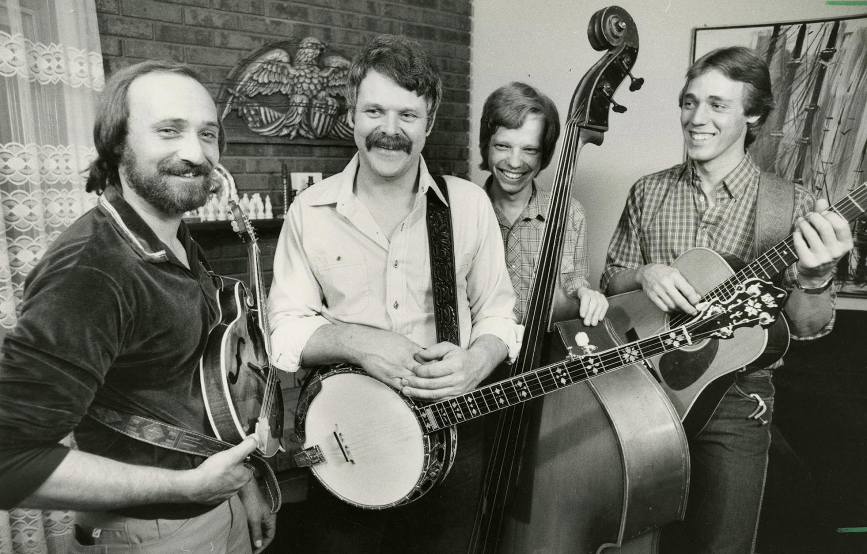 Four casually dressed men in living room by fireside mantle, from left one with guitar, second …