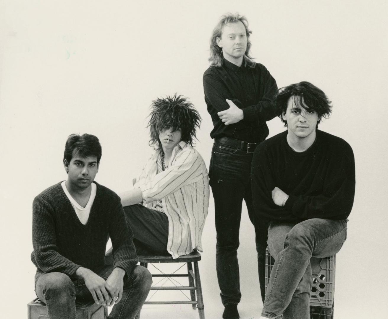 Four young men variously dressed in black sweaters, shirts and jeans, three sitting and one sta…