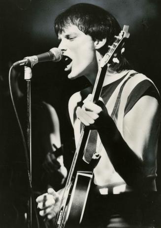 Young man sings into microphone on stand while he plays a bass guitar on darkened stage. 