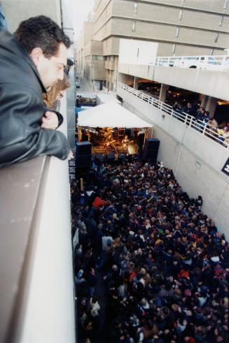 Man leans over ledge looks down on a wall to wall crowd in an alley in front of a stage where G…