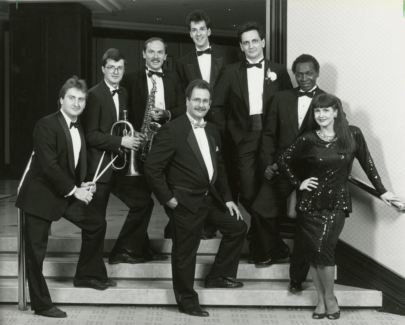 Full body image on a few stairs,a man is surrounded by six other men and a woman on the right. …