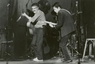 Full body shot. Two young men playing guitars seen sideways on darkened stage.