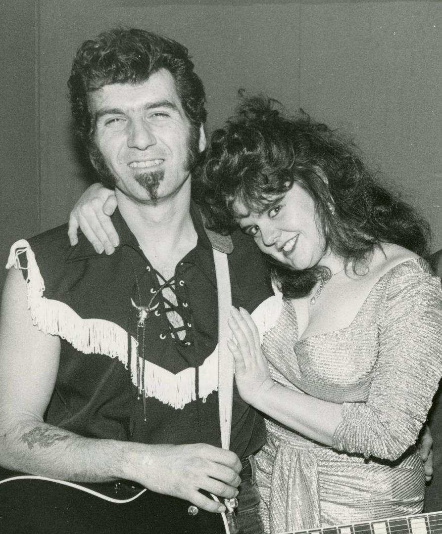 Man on left with goatee and fringed sleeveless shirt is hugged by woman on his right with long …