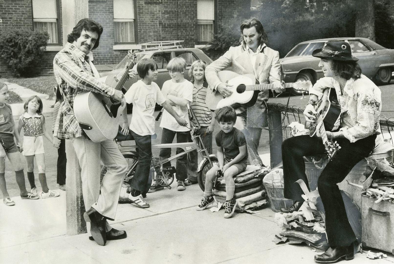 Daytime on street corner, three men in sport coats and boots playing guitars and singing with a…