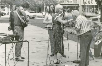 Summer day, outside on sidewalk, an older man kisses the hand of a woman holding violin. Behind…